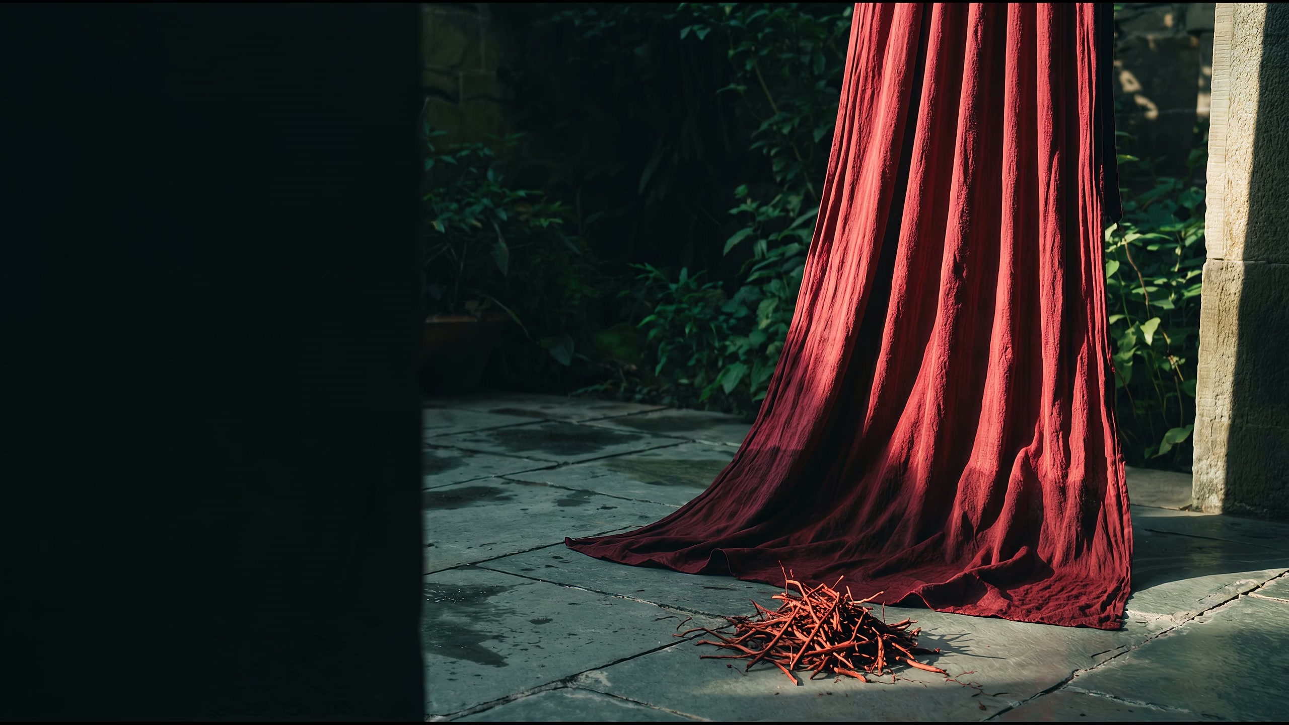 A length of crimson cotton hanging in a shaded stone courtyard, with madder roots heaped on the wet flagstones beneath it.